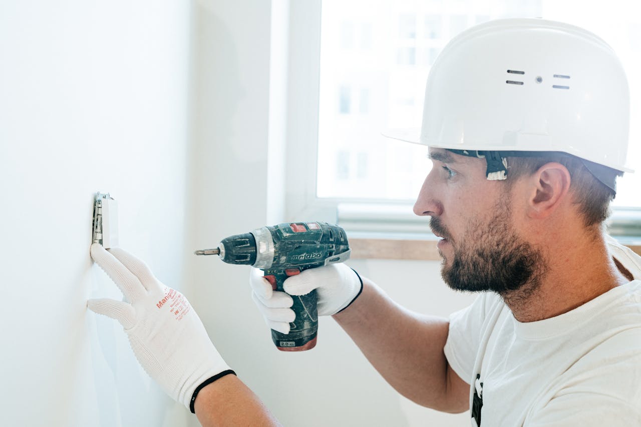 Side view of a construction worker using a drill on a wall, wearing safety gear.